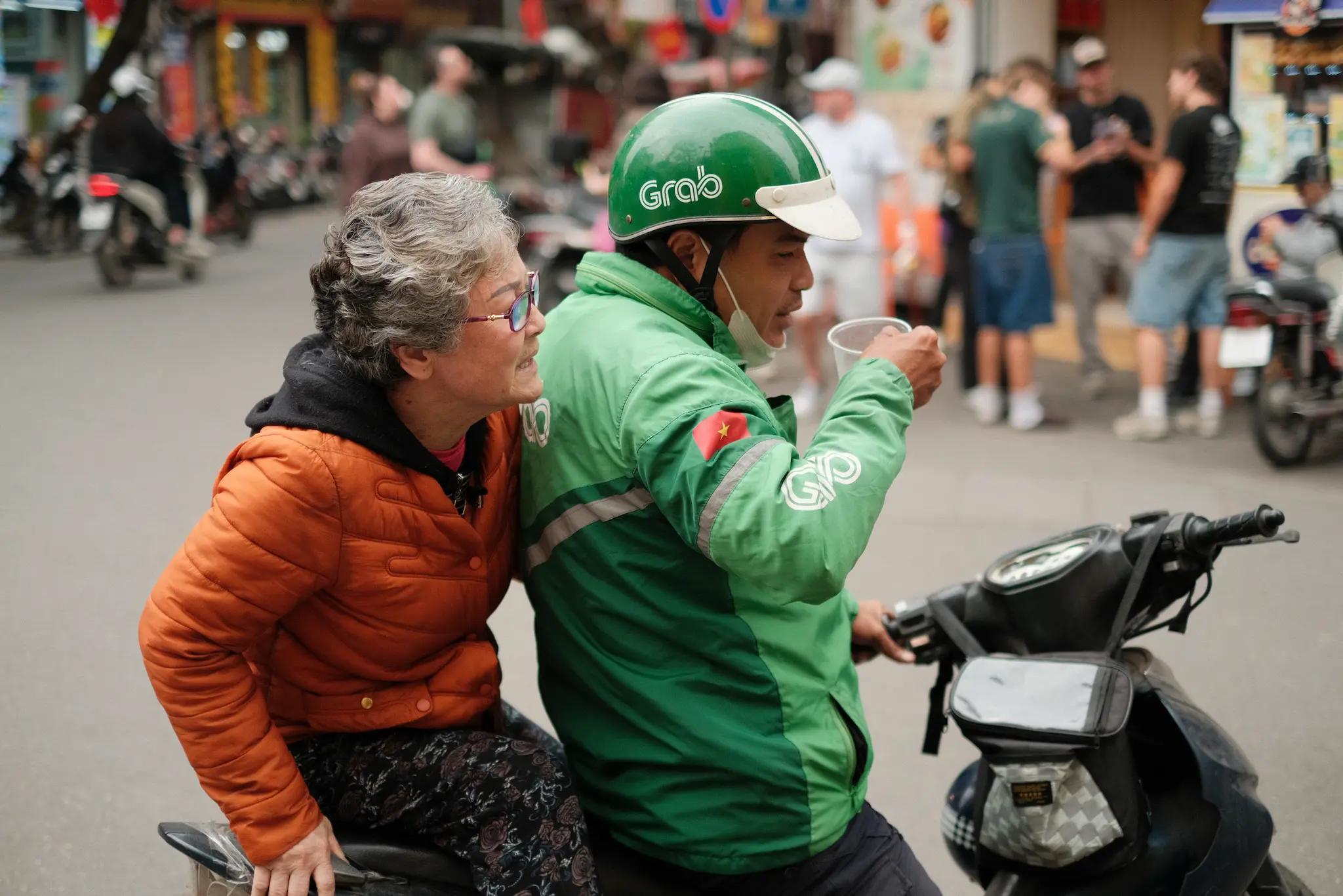chauffeur Grab en veste verte et passagère âgée assis sur un scooter dans une rue animée de Hanoï, photographie de rue Fujifilm